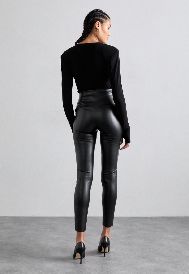 Schwarzes Langarmoberteil, kombiniert mit hochgeschnittenen schwarzen Leggings aus Kunstleder, die eine glatte Textur und einen figurbetonten Schnitt aufweisen, akzentuiert mit hohen Schuhen.