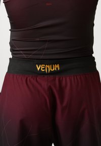 Μαρόν αθλητικά σορτς με μαύρη ζώνη, διαθέτοντας το χρυσό λογότυπο "VENUM". Λείος ύφασμα με υποβαθμισμένο σχέδιο και ορατές λεπτομέρειες ραφής.