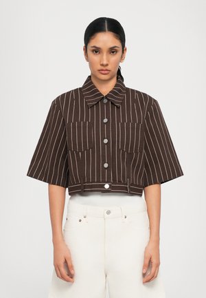Brunstripete cropped jakke med krage, to frontlommer og sølvknapper. Stoffet ser teksturert ut, kombinert med hvite bukser.
