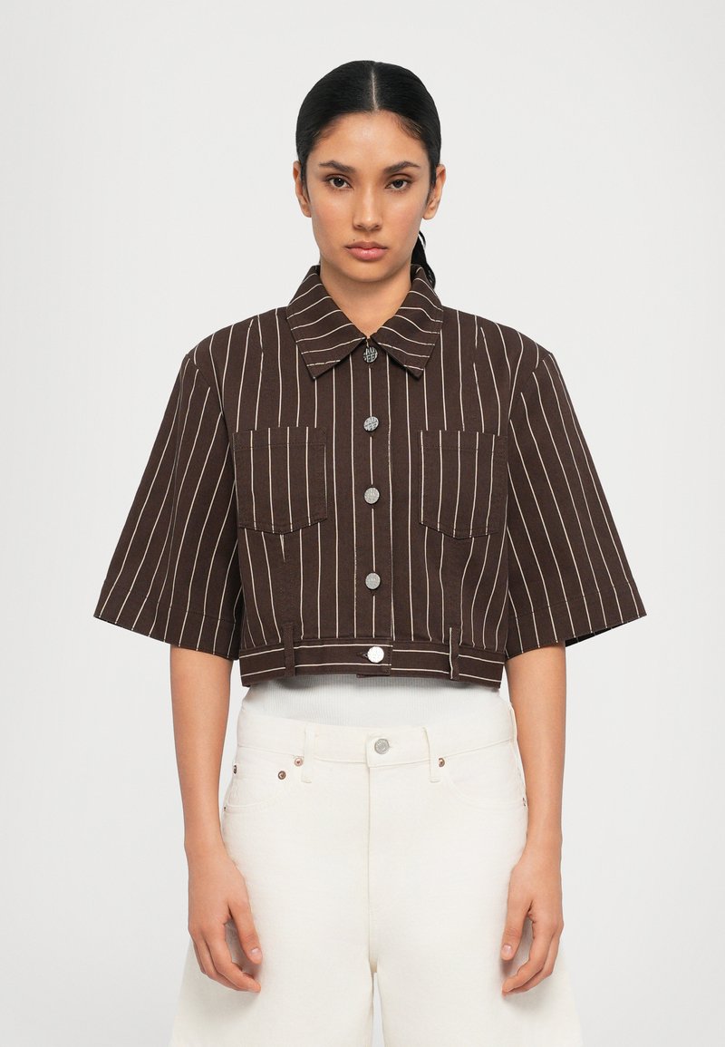 Brown striped cropped jacket with a collar, two front pockets, and silver buttons. The fabric appears textured, paired with white pants.