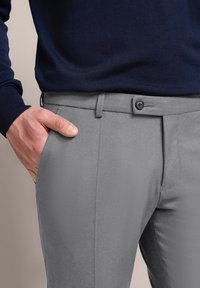 Graue Hose mit glatter Textur, taillierter Passform und Reißverschluss. Sie verfügt über seitliche Taschen und einen Knopfverschluss und wird mit einem dunkelblauen Pullover kombiniert.