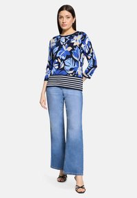 Femme portant un haut bleu à motifs floraux avec un ourlet rayé, un jean large bleu clair, et des talons noirs ouverts, debout devant un fond blanc.