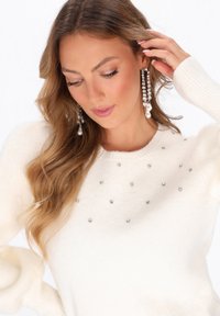 Maglione bianco a maglia con scollo rotondo e piccole pietre brillanti sparse sul davanti. Maniche lunghe con polsini a costine.