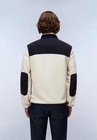 Person med krøllet brunt hår iført en creme- og sort jakke med sorte albuelapper, vist bagfra mod en ensfarvet baggrund.