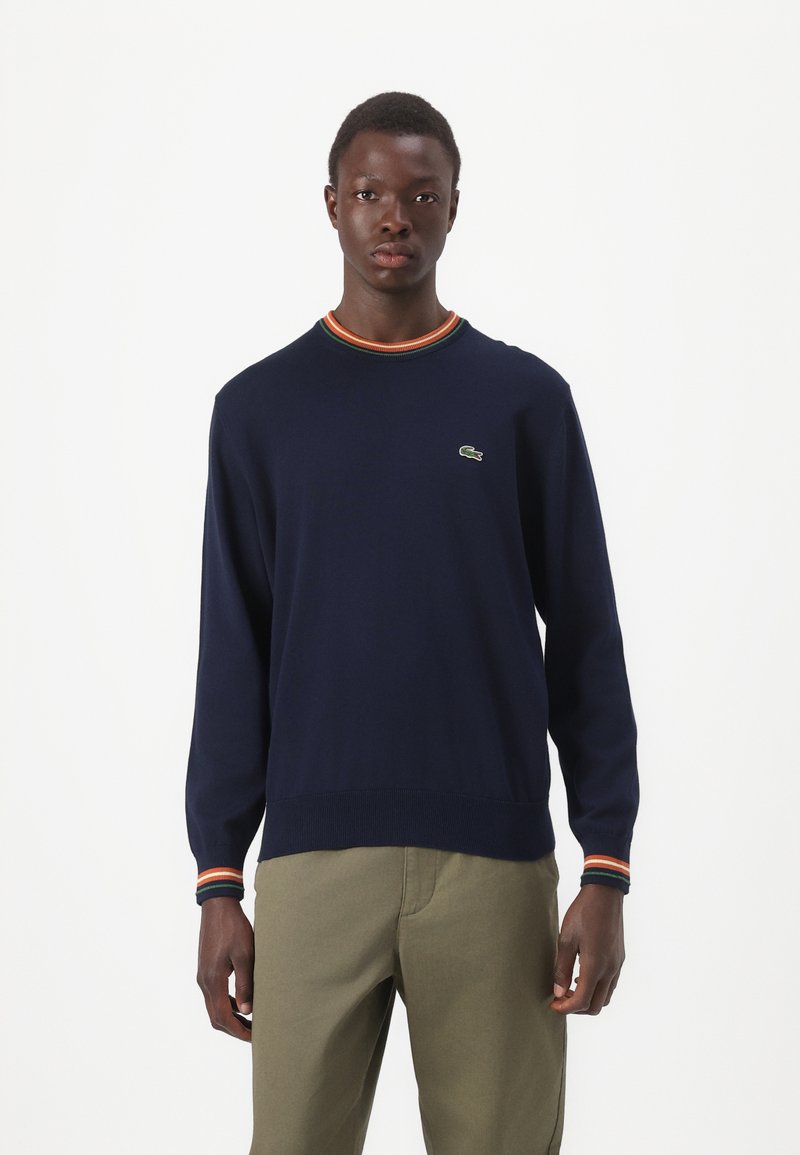 Marineblauwe trui met lange mouwen en geribbelde halslijn, met oranje, rode en groene strepen. Lacoste-logo op de borst; gebreide textuur.