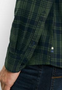 Grün und marineblau karierter Flanellhemdärmel mit Knöpfen, mit einer glatten Textur und einem kleinen Logoschildchen am Saum.