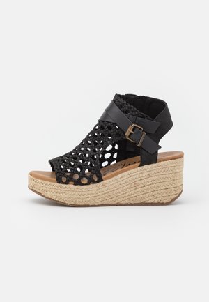 Sandale compensée tressée noire avec un design en crochet, des accents en cuir et une base en liège enveloppée de jute, dotée d'un détail de bride à boucle.