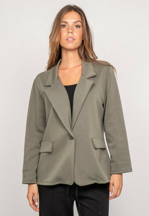 Femme portant un blazer vert olive ample sur un haut noir et un pantalon noir, debout devant un fond clair uni.