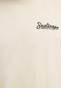 Sweat-shirt de couleur crème avec un col rond côtelé et un logo noir "Soulscape" sur la poitrine. Le tissu a une apparence douce et texturée.