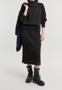 Personne portant une jupe en maille côtelée sombre et un pull assorti, tenant une veste bleu marine sur l'épaule et portant des bottes de combat noires à lacets.