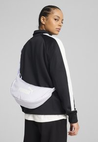 Poseta de culoare lavandă deschisă, cu o suprafa�ță texturată, prezentând logo-ul "PUMA" și un design curbat, purtată peste o jachetă de trening neagră.