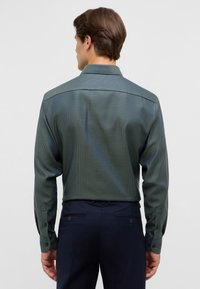 Man die met zijn rug naar de camera staat, gekleed in een groen gestreept hemd met lange mouwen en donkerblauwe broek tegen een effen achtergrond.