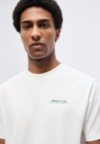 Νεαρός άνδρας με σγουρά μαλλιά που φοράει λευκό T-shirt με πράσινο κείμενο «JUICE IT UP Marc O'Polo Denim» σε απλό φόντο.