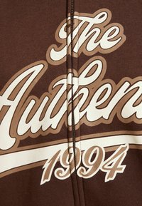 Hnědá mikina na zip s velkým krémovým a světle hnědým textem: "The Authentic 1994." Měkká tkanina s volným designem.
