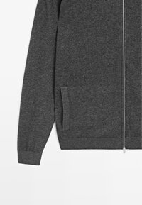 Grå zip-up tröja i mjukt, strukturerat tyg. Har långa ärmar, en framficka och en silverfärgad dragkedja som går ner i mitten.