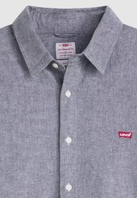 Camicia grigia in chambray con colletto classico, bottoni bianchi e un piccolo logo rosso Levi's sul lato sinistro del petto. Finitura in tessuto testurizzato.