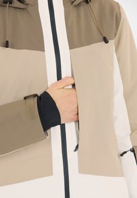 Giacca beige e bianca con cappuccio, caratterizzata da un design a blocchi di colore e una cerniera verticale. Una mano sta raggiungendo una tasca laterale.