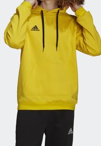 Personne portant un sweat à capuche jaune Adidas avec des cordons noirs et un pantalon Adidas noir, les mains ajustant la capuche.