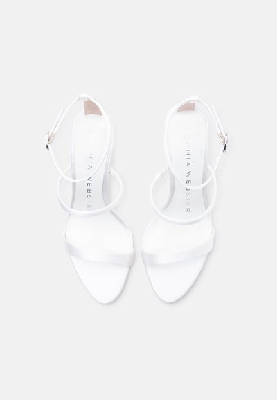 Λευκά ψηλοτάκουνες σανδάλια με λουρίδες, με αγκράφες στον αστράγαλο και λεπτές μπροστινές λουρίδες, απεικονιζόμενα από πάνω σε απλό φόντο.