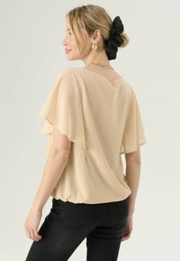Beige chiffon top met vlindermouwen, losse pasvorm, diepe rug, gedragen met zwarte skinny jeans. Haar opgestoken met een zwarte scrunchie.