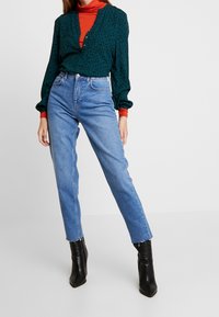 Blusa verde con estampado de leopardo negro, de manga larga y con escote en V. Combinada con jeans de tiro alto en un tono azul claro y botas negras hasta el tobillo.