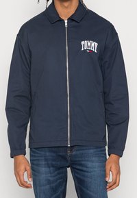 Marineblå zip-jacka i ett slätt tyg, med en logotyp på vänster bröst, elastiska cuffs och sidofickor.