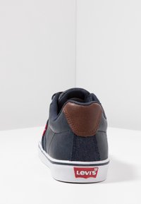 Baskets bleu marine avec un accent en cuir marron au niveau du talon, semelle blanche, détail du logo rouge, et empeigne en textile. Texture lisse, design sportif.