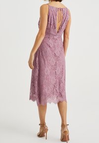 Spetsklänning i ljusviolett med vågig fåll, V-formad rygg och knutna band. Tyget har blomsterpattern och en figurnära silhuett.