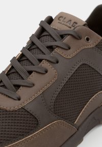 Brown athletic shoe featuring a mesh upper, contrasting smooth accents, and flat laces. Logo embossed on the tongue. Textured materials visible.