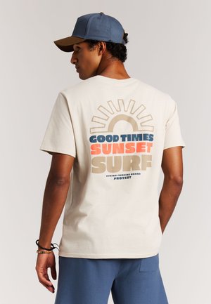 Crèmekleurig T-shirt met een zonafbeelding en de tekst "Good Times Sunset Surf" in blauw, oranje en beige; gecombineerd met een blauw-bruine pet.