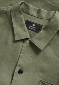 Camicia verde con colletto, in tessuto texturizzato, un bottone nero e un'etichetta con cuciture dorate che indica "Matinique Heritage Apparel."