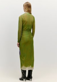 Grünes, leicht transparentes Kleid mit langen Ärmeln und Raffung auf der Rückseite. Verfügt über einen fließenden Rock und einen kontrastierenden Saum. Kombiniert mit schwarzen knielangen Stiefeln.