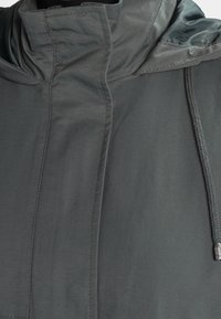 Gros plan sur une veste à capuche gris foncé avec fermeture éclair frontale cachée, coutures visibles et cordon de serrage avec embouts en métal sur le côté droit.