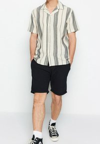 Man met een kortemouwen beige shirt met verticale grijze strepen, zwarte shorts, witte sokken en zwarte hoge sneakers.
