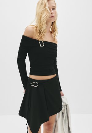 Top nero off-the-shoulder in morbido tessuto con un accento argento. Abbinato a una gonna asimmetrica nera, anch'essa con un dettaglio argentato.