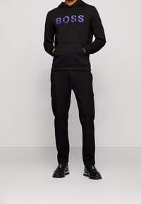 Černá mikina s kapucí s fialovým logem "BOSS" a s klokaní kapsou. Kombinováno s černými cargo kalhotami a černými teniskami.