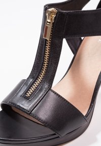 Sandal en cuir noir avec un bout ouvert, de larges lanières et un détail en zip doré sur le devant. Texture lisse avec un léger éclat.