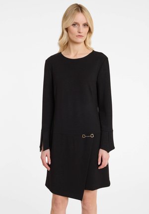 Zwarte jurk van gladde stof, met lange mouwen, een ronde halslijn en een asymmetrische zoom met gouden kettingdetail op de taille.