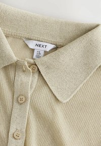Beige ribattu poloshirt dengan kerah, tiga butang, dan label saiz yang menunjukkan UK 8, EU 36, US 4, dibuat di Turki.