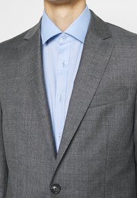 Szary marynarka o fakturze z wyciętymi klapami i pojedynczym guzikiem, zestawiona z jasnoniebieską koszulą z kołnierzykiem i mankietami zapinanymi na guziki.