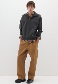 Pullover grigio con colletto a zip, caratterizzato da una tasca frontale, sovrapposto a una camicia a quadri. Pantaloni cargo marroni con tasche laterali, scarpe nere.