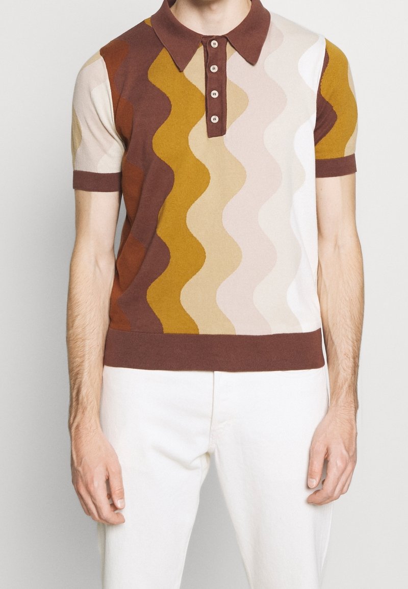 Muž v polo tričku s krátkým rukávem s vlnitými svislými pruhy v hnědé, hořčicové, béžové a bílé barvě, spárovaný s bílými kalhotami.