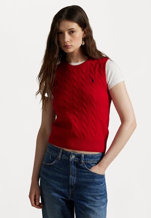Rød kabelstrikket utenermet genser med ribbestrikket kant, lagt over en hvit kortermet skjorte, kombinert med blå denim jeans.