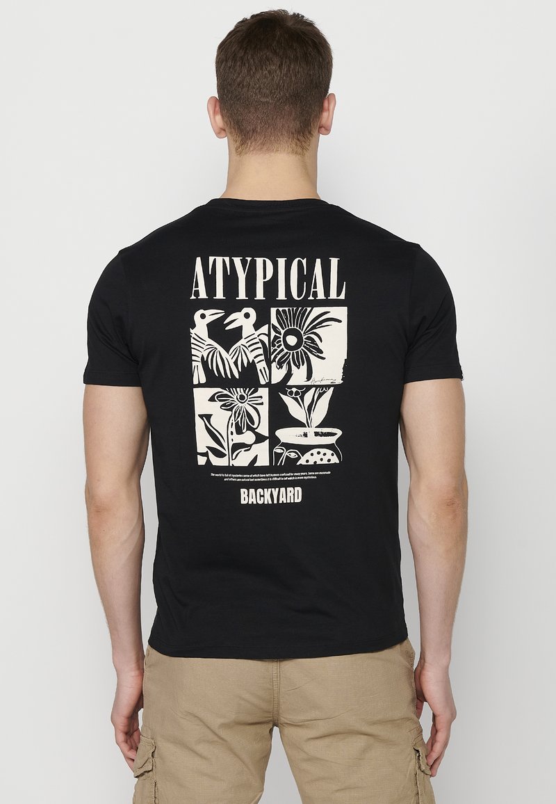 Melnas kokvilnas T-kreklas ar baltu grafisko dizainu aizmugurē, kas ietver stilizētus putnus un ziedu rakstus, ar tekstu "ATYPICAL" un "BACKYARD."