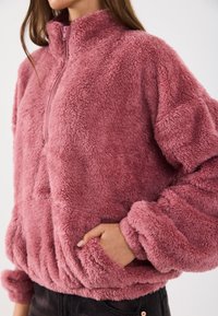 Rozā, pūkaina džemperis, izgatavots no mīksta materiāla ar augstu apkakli, ar zīmuļu priekšpusi un sānu kabatām. Iekļautas elastīgas aproces un apakšējā mala.