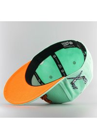 Vihreä minttulippis, jossa on oranssi lippa. Sisältää mustan vuorauksen ja brodeeratun kuvion, joka esittää sakset ja savua.