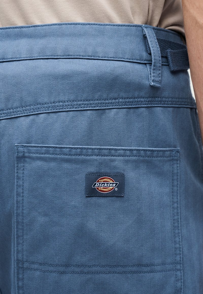 Blue work pants with a back pocket featuring the Dickies logo patch, worn by a person in a beige shirt.