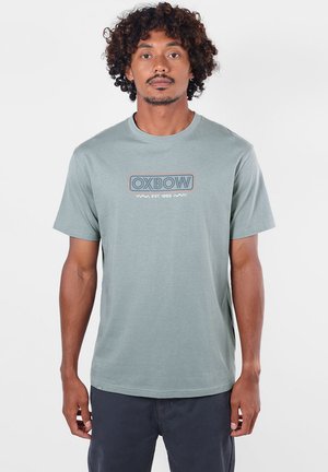 Homme aux cheveux bouclés portant un t-shirt Oxbow vert clair et un pantalon foncé, debout face à l'avant avec une expression neutre.