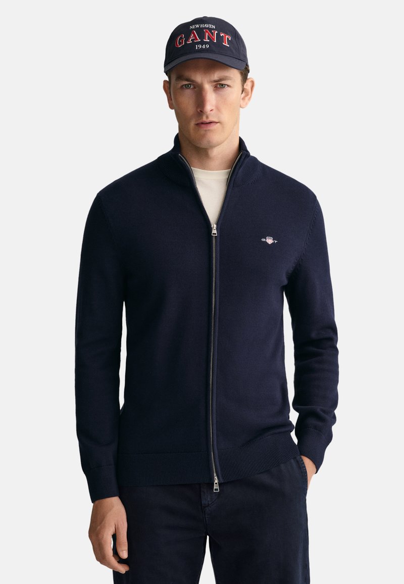 Pull à fermeture éclair bleu marine avec un col haut, fabriqué en tissu tricoté doux ; présente un petit logo sur la poitrine gauche et des poignets côtelés. Casquette bleu marine avec logo.