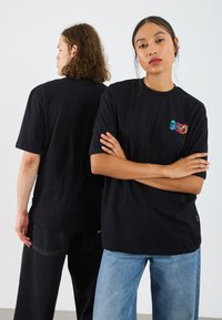 Black cotton t-shirt with short sleeves, featuring a small colorful graphic on the chest. The fit is relaxed, with a straight cut.